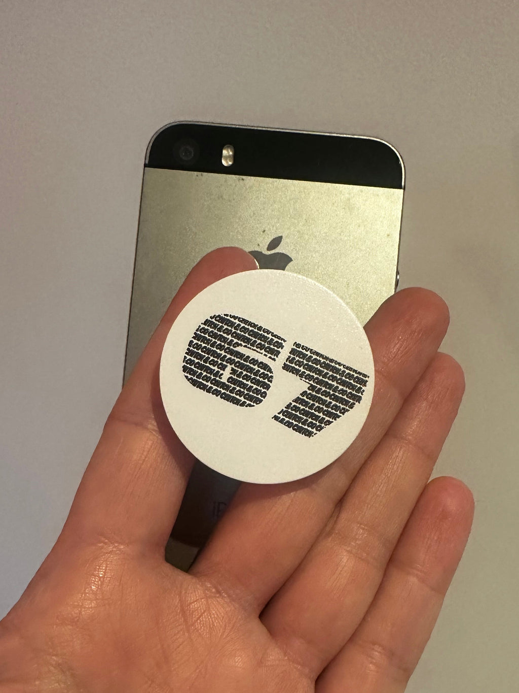Hand holding a round white sticker with '67' logo on a silver phone