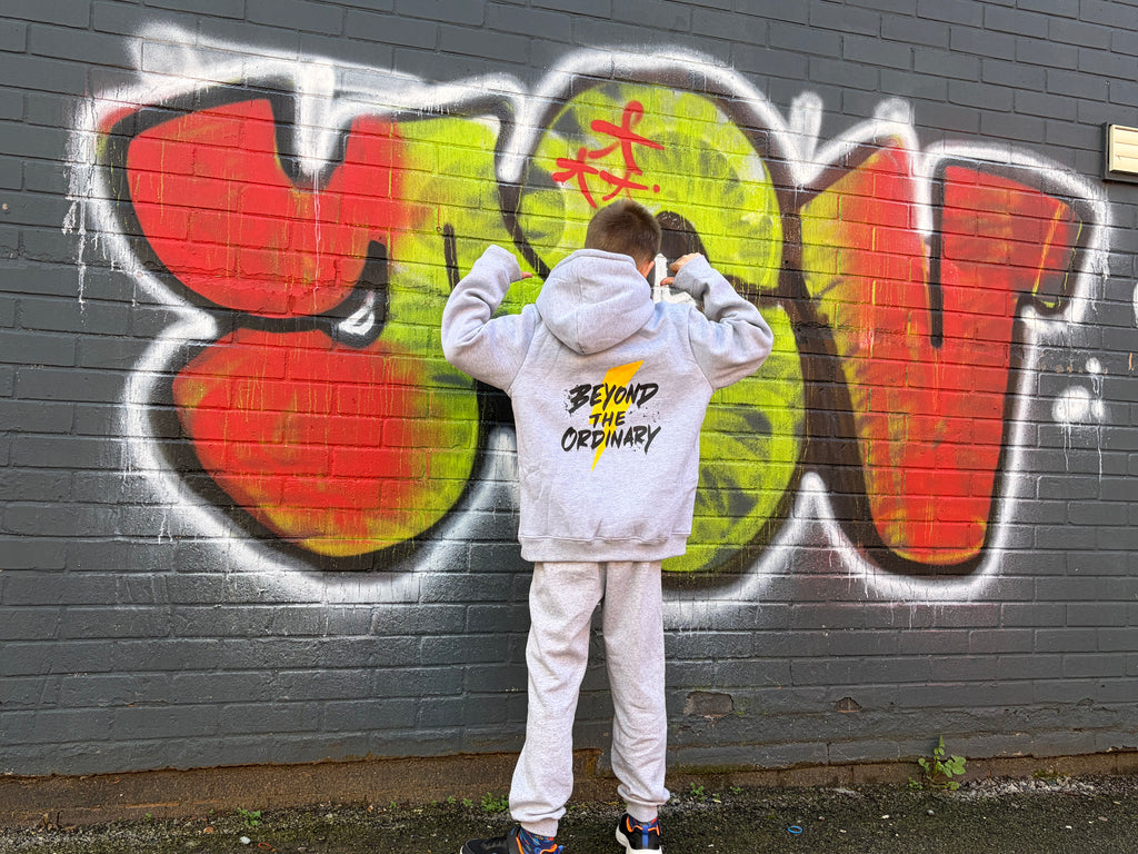 Person wearing a hoodie with 'Beyond the Ordinary' text in front of colorful graffiti on a wall.