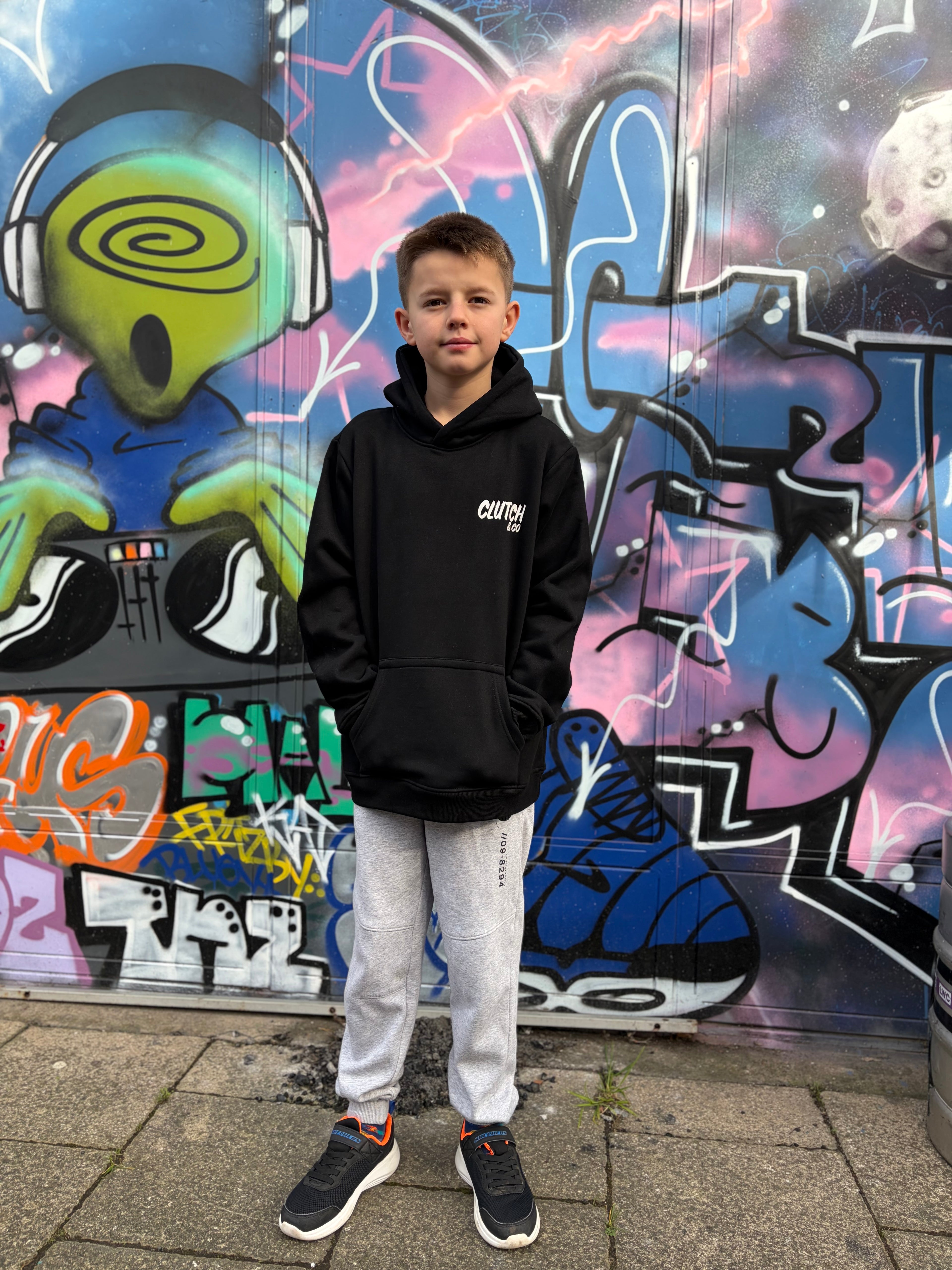 Young boy wearing a black hoodie with text, standing in front of a colorful graffiti wall.