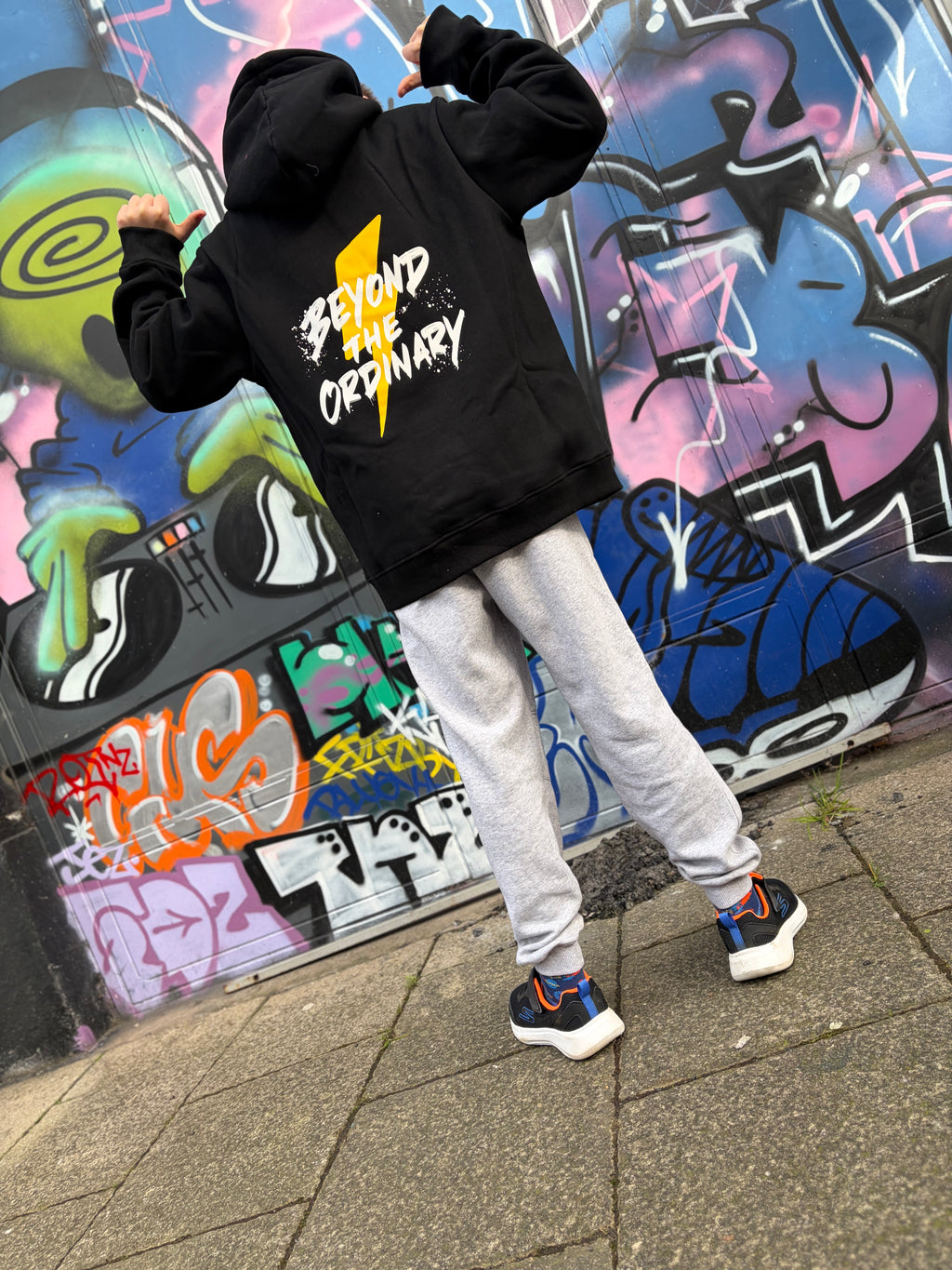 Person wearing a black hoodie with 'Beyond the Ordinary' text in front of a colorful graffiti wall.