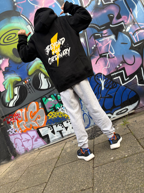 Person wearing a black hoodie with 'Beyond the Ordinary' text in front of a colorful graffiti wall.