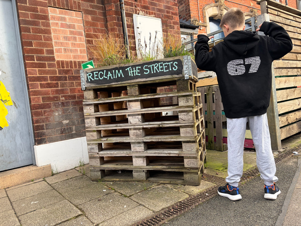 Person wearing a black hoodie with '67' on it standing next to a wooden pallet structure with 'Reclaim the Streets' sign.
