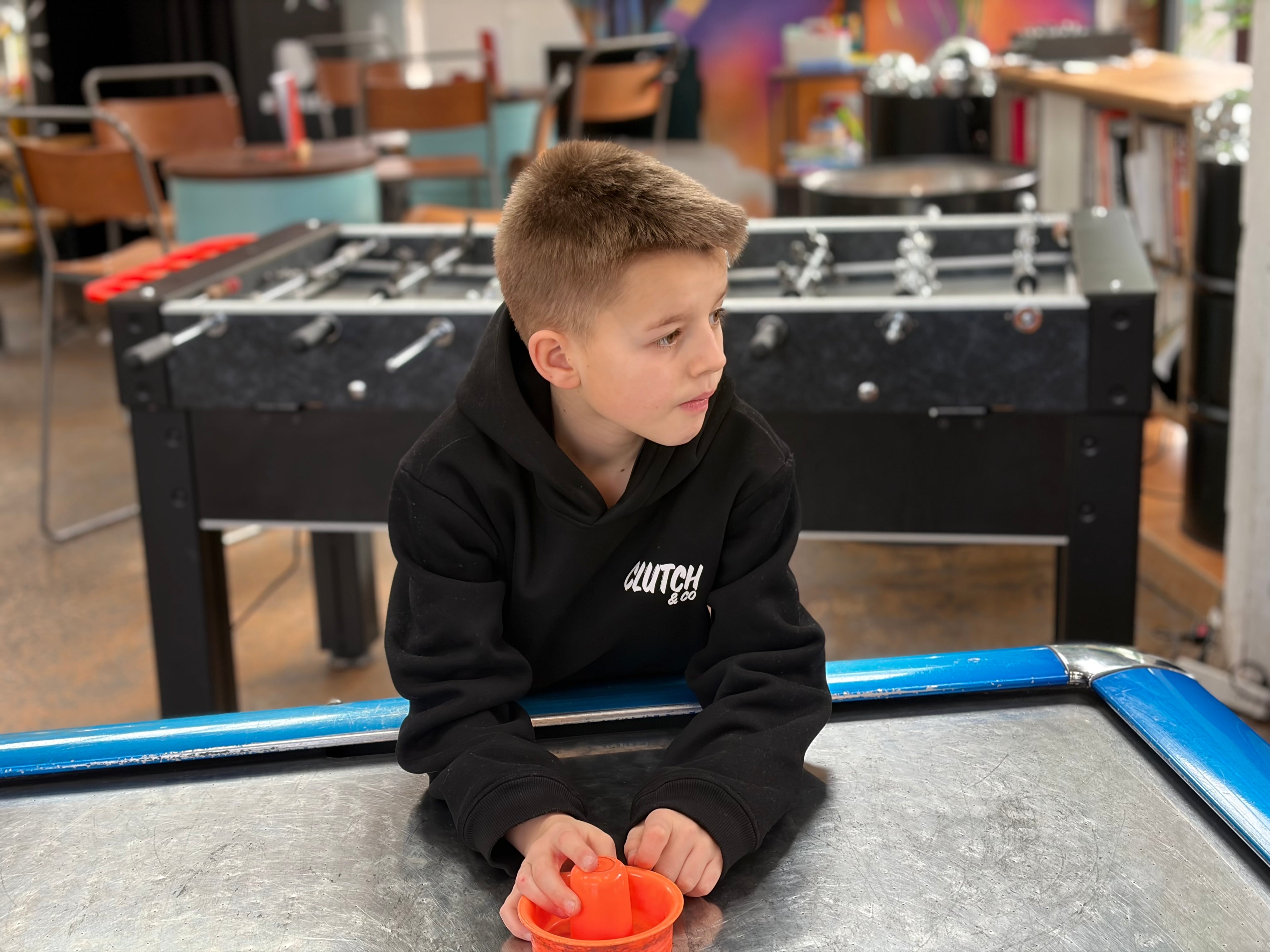 Young boy in a black hoodie sitting at a foosball table in an indoor setting.