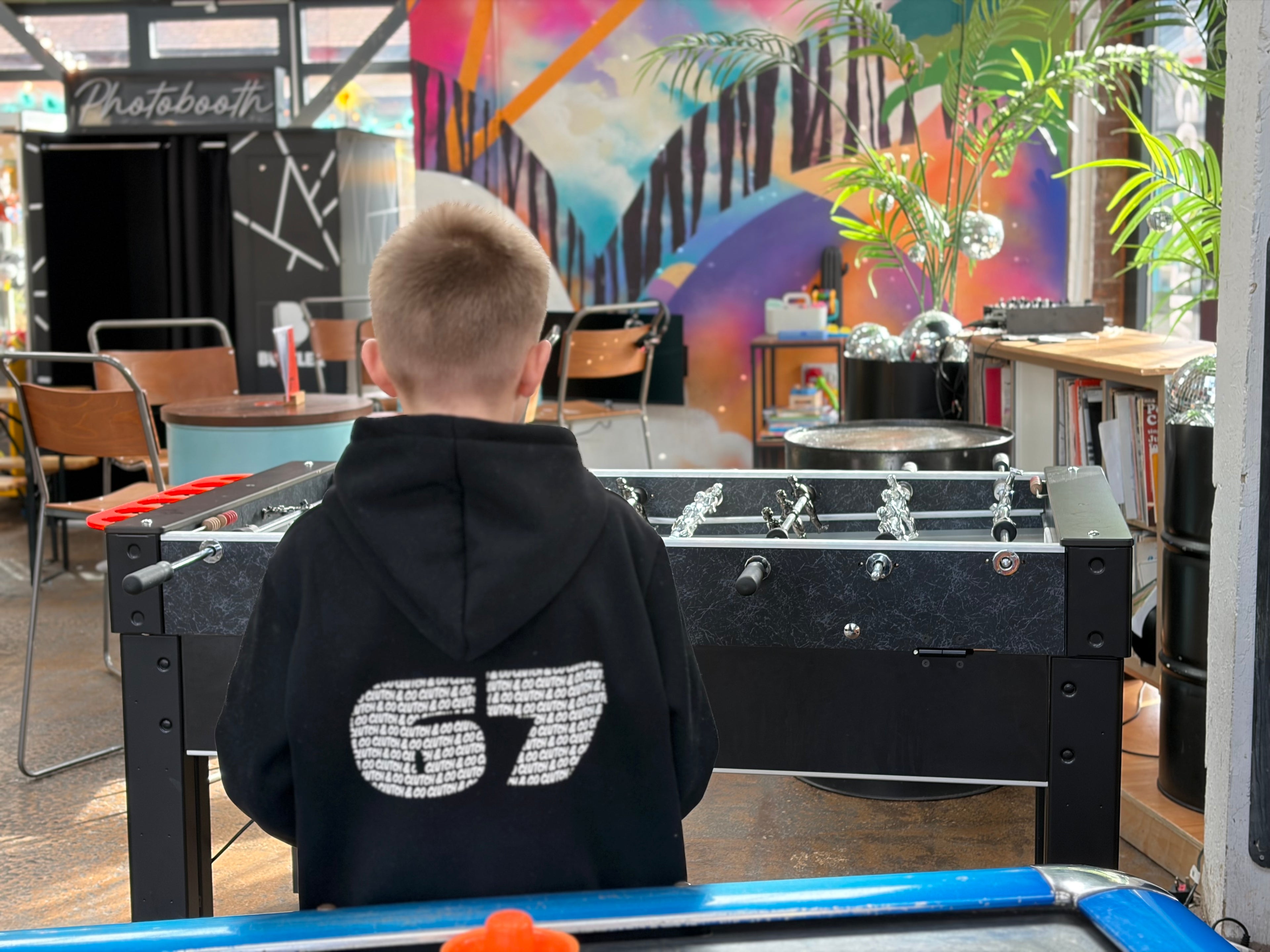 Child wearing a black hoodie with '67' on it, standing in front of a foosball table in a colorful indoor setting.